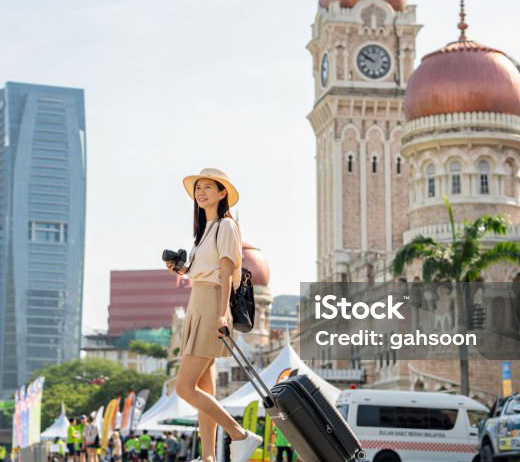 female tourist walking around sultan abdul samad building in kuala lumpur, holding camera and suitcase wearing hat and backpack